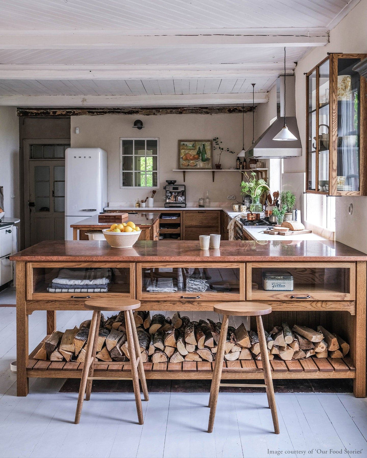 A straight-on view of a Scandinavian-style oak kitchen with an island peninsula and two simple oak bar stools with oval-shaped seats and brass footrails. TEST