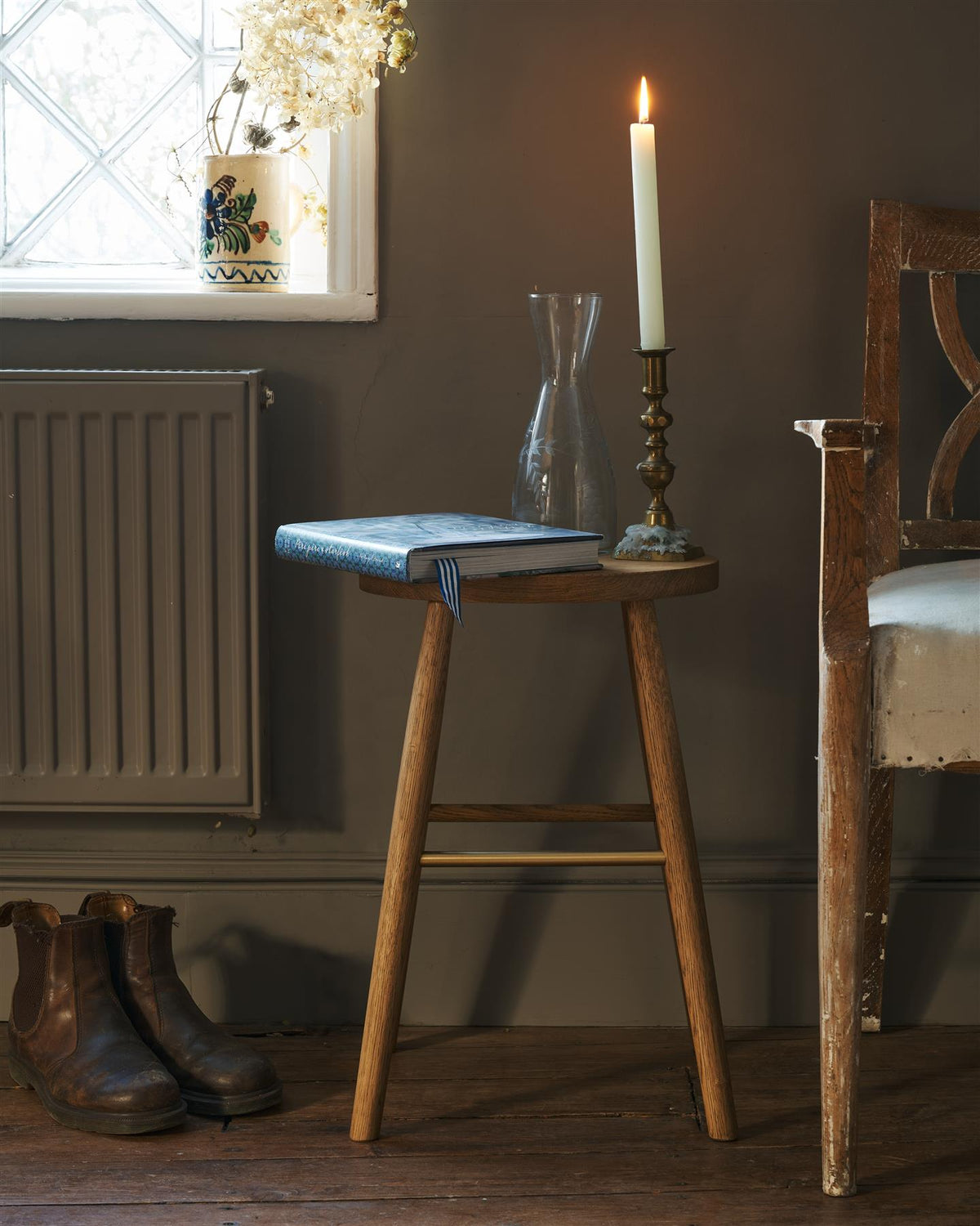 A straight-on view of a simple oak milking stool with an oval-shaped seat and a brass footrail in a dark room. A hardback book and a lit pillar candle are placed ontop of the stool. TEST