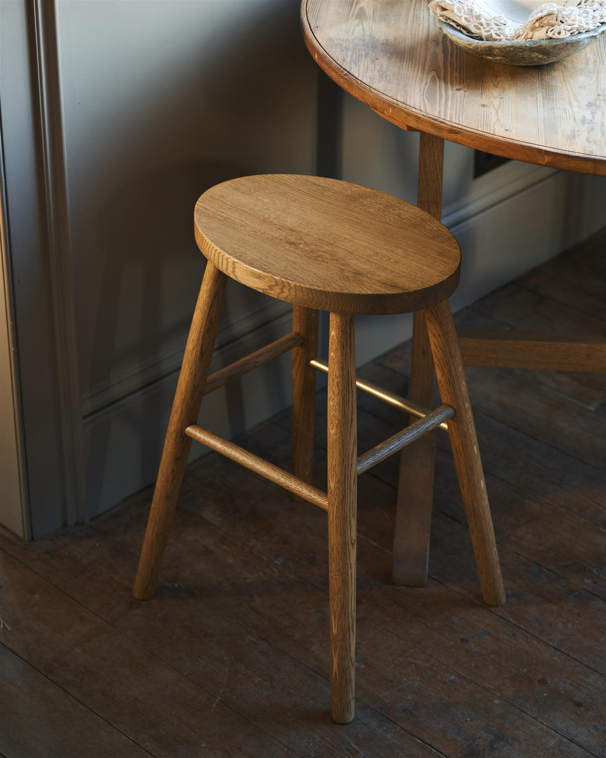 An oblique view of a simple oak milking stool with an oval-shaped seat and a brass footrail. TEST