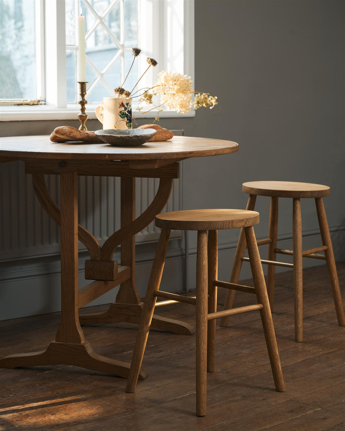 An oblique view of two simple oak milking stools with oval-shaped seats and brass footrails by a round pine table in a dark room. TEST