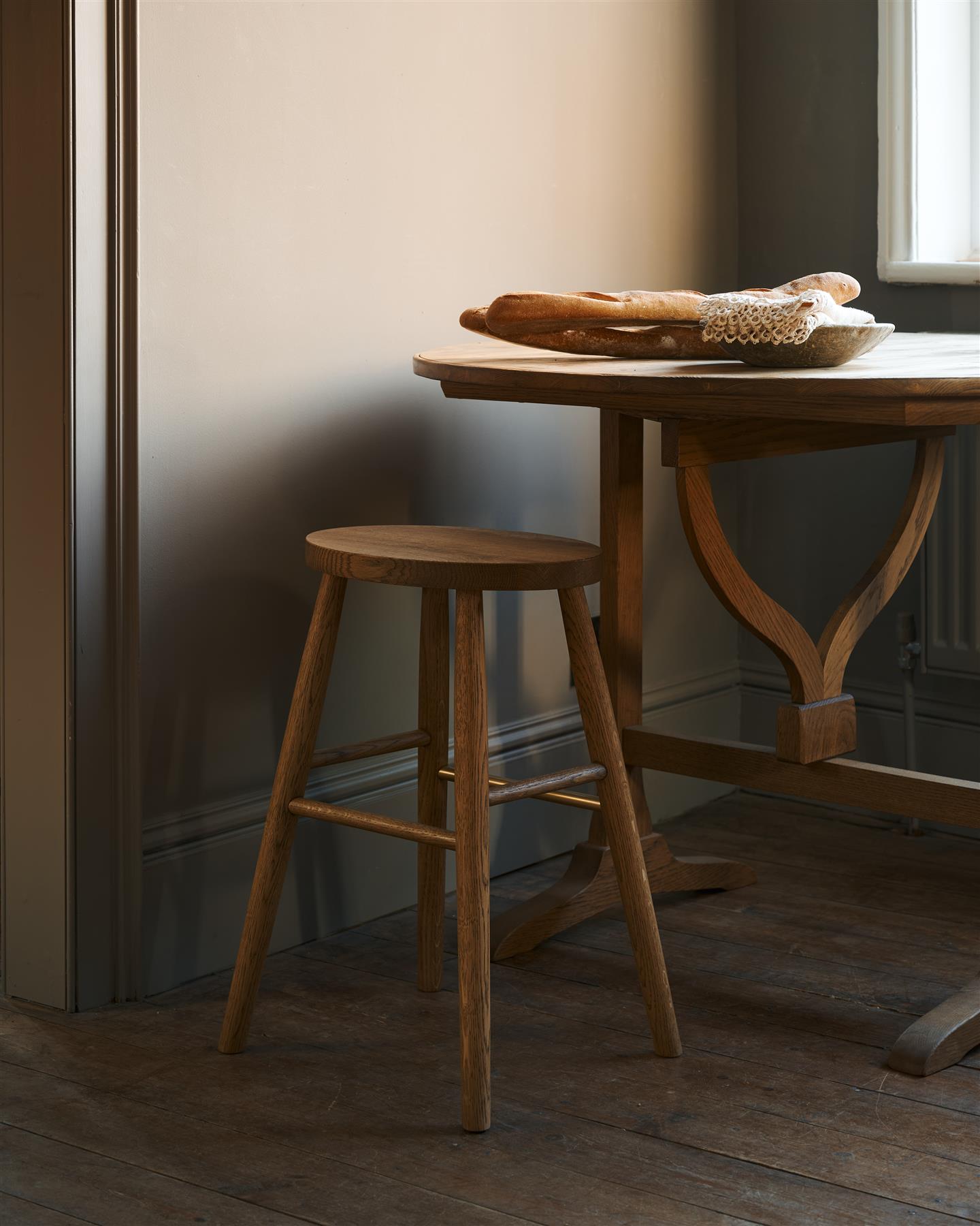 An oblique view of a simple oak milking stool with an oval-shaped seat and a brass footrail by a round pine table in a dark room.