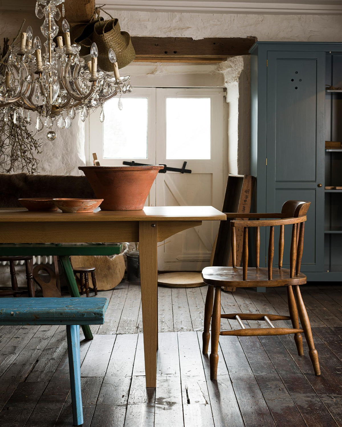 A straight-on view of the end of a mid-century modern-style oak dining table in a rustic cottage with dark worn floorboards and white brick walls. TEST