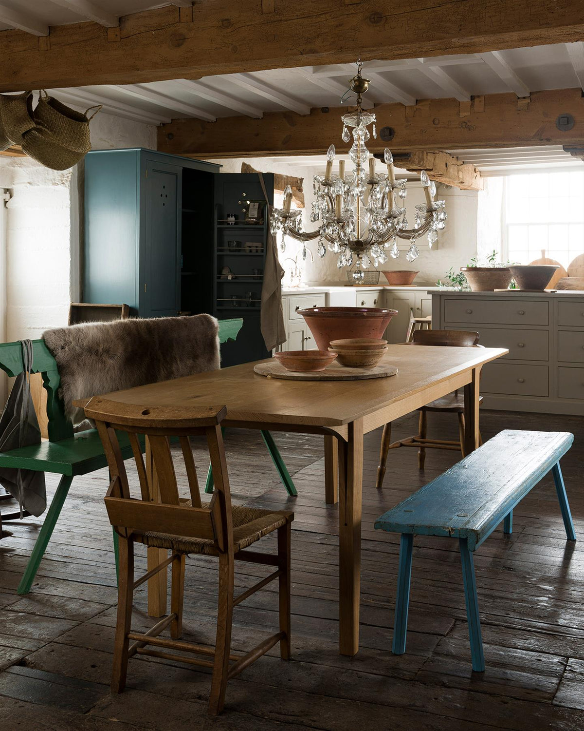 An oblique view of a mid-century modern-style oak dining table in a rustic cottage with dark worn floorboards and white brick walls. TEST