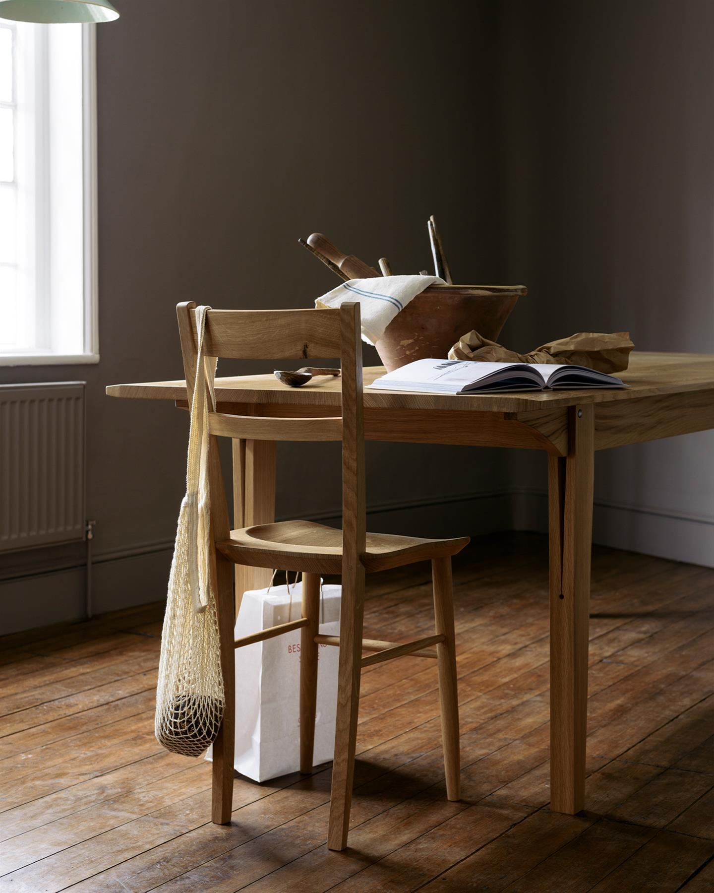 An oblique view of mid-century-style dining chair at the end of a mid-century modern-style oak dining table in a dark room with wooden floorboards.
