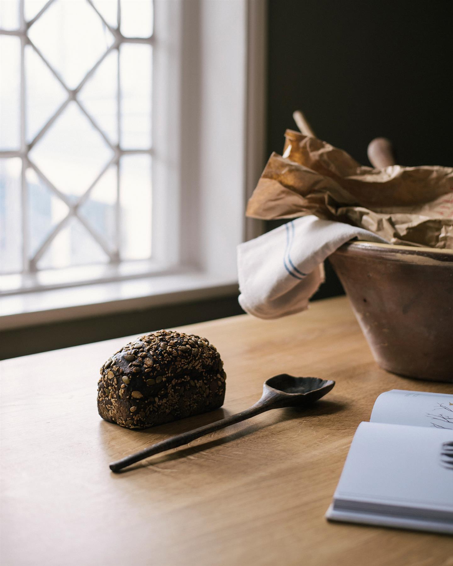 An oblique view of an oak tabletop with a large ceramic bowl and seeded bread in front of a Victorian leaded window.