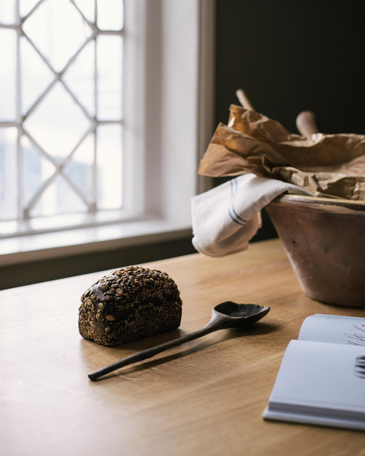 An oblique view of an oak tabletop with a large ceramic bowl and seeded bread in front of a Victorian leaded window. TEST