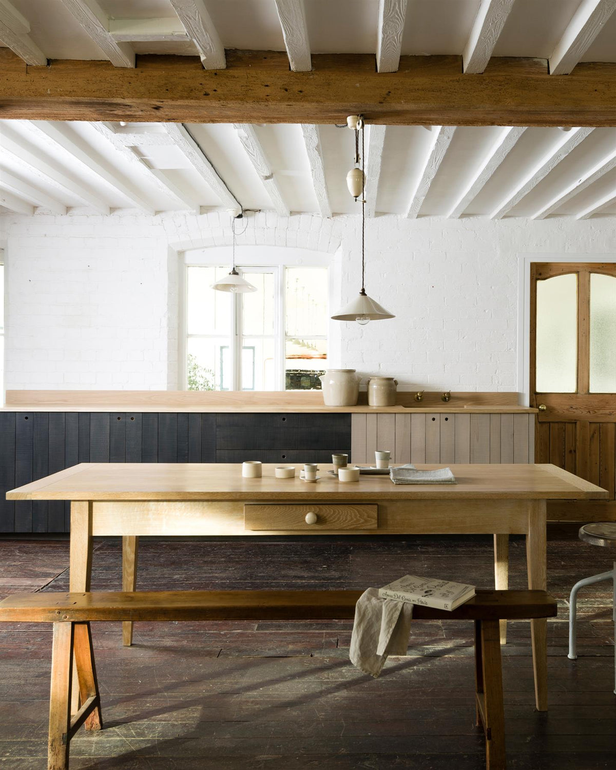 A straight-on view of a Shaker-style oak dining table with breadboard ends, in a cottage room with dark worn floorboards, white brick walls and beamed ceilings. TEST