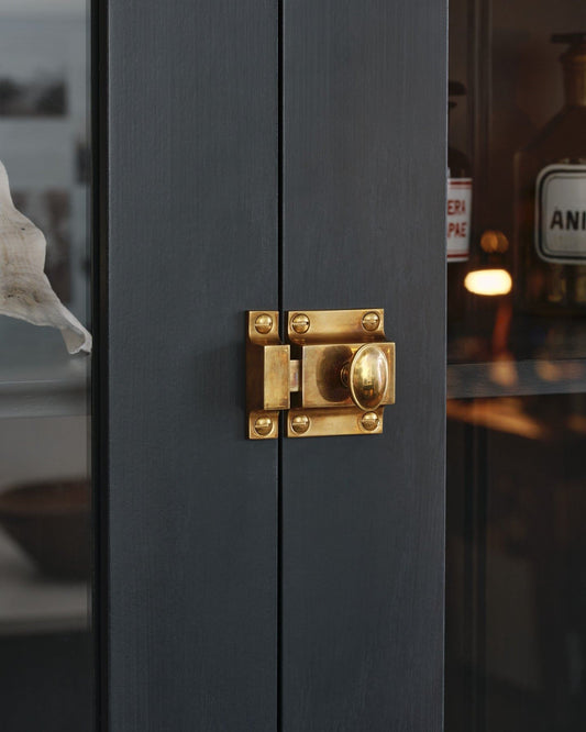 An oblique view of a classic-style square cupboard catch with an ornate oval-shaped knob in a warm burnished brass finish fitted to dark blue painted glazed cupboard doors.