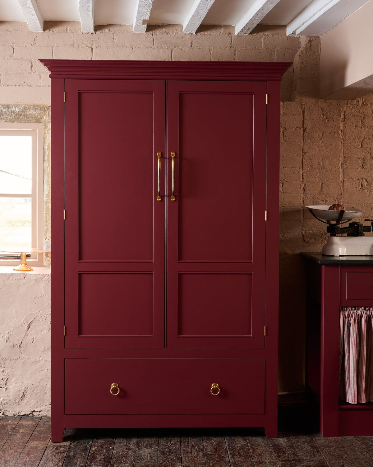 A tall red painted Georgian-style pantry cupboard with double doors and a bottom drawer, fitted with Victorian-inspired ring pull handles in a warm burnished brass finish. TEST