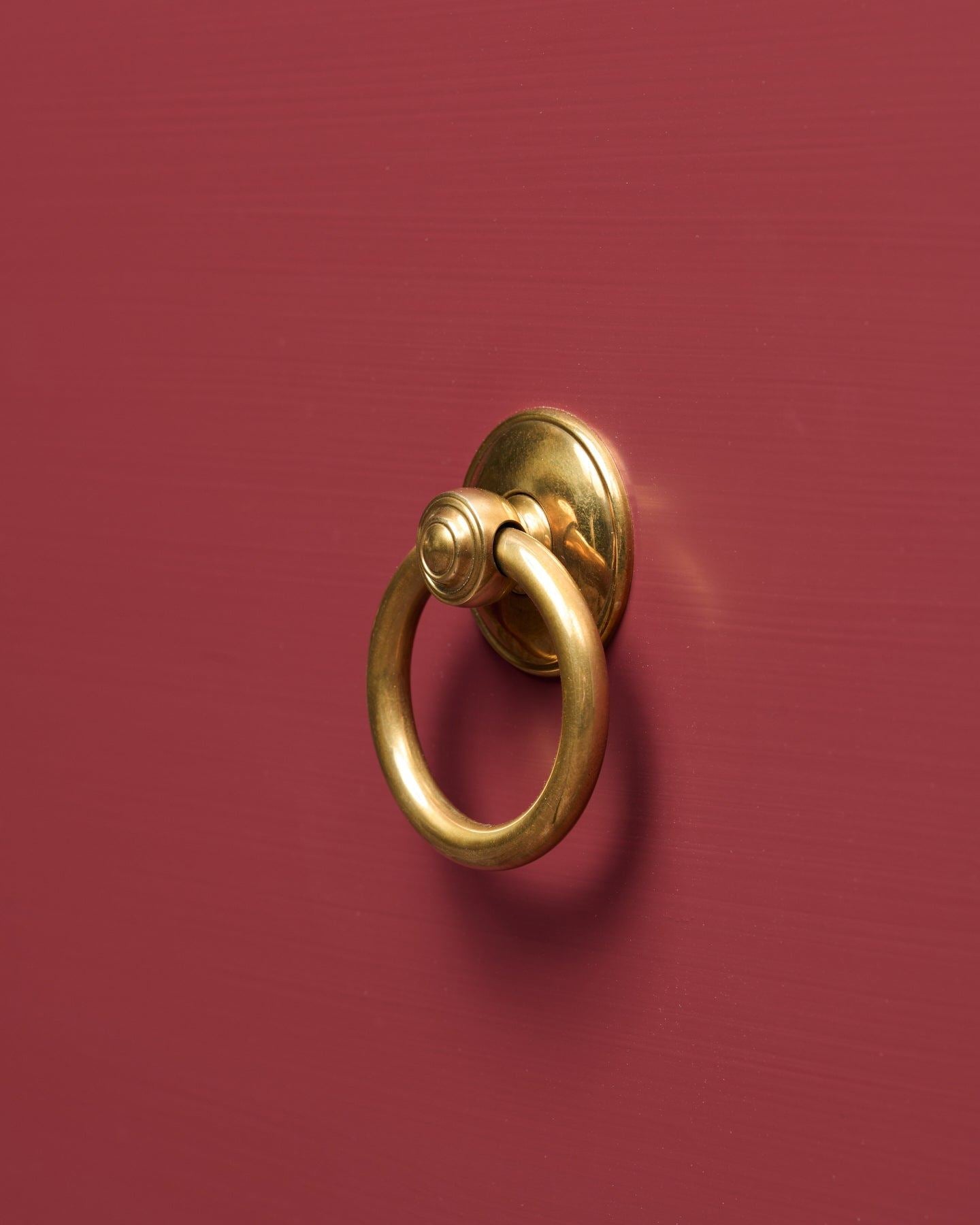 An oblique view of a Victorian-inspired ring pull handle in a warm burnished brass finish, fitted to a red painted cupboard drawer.