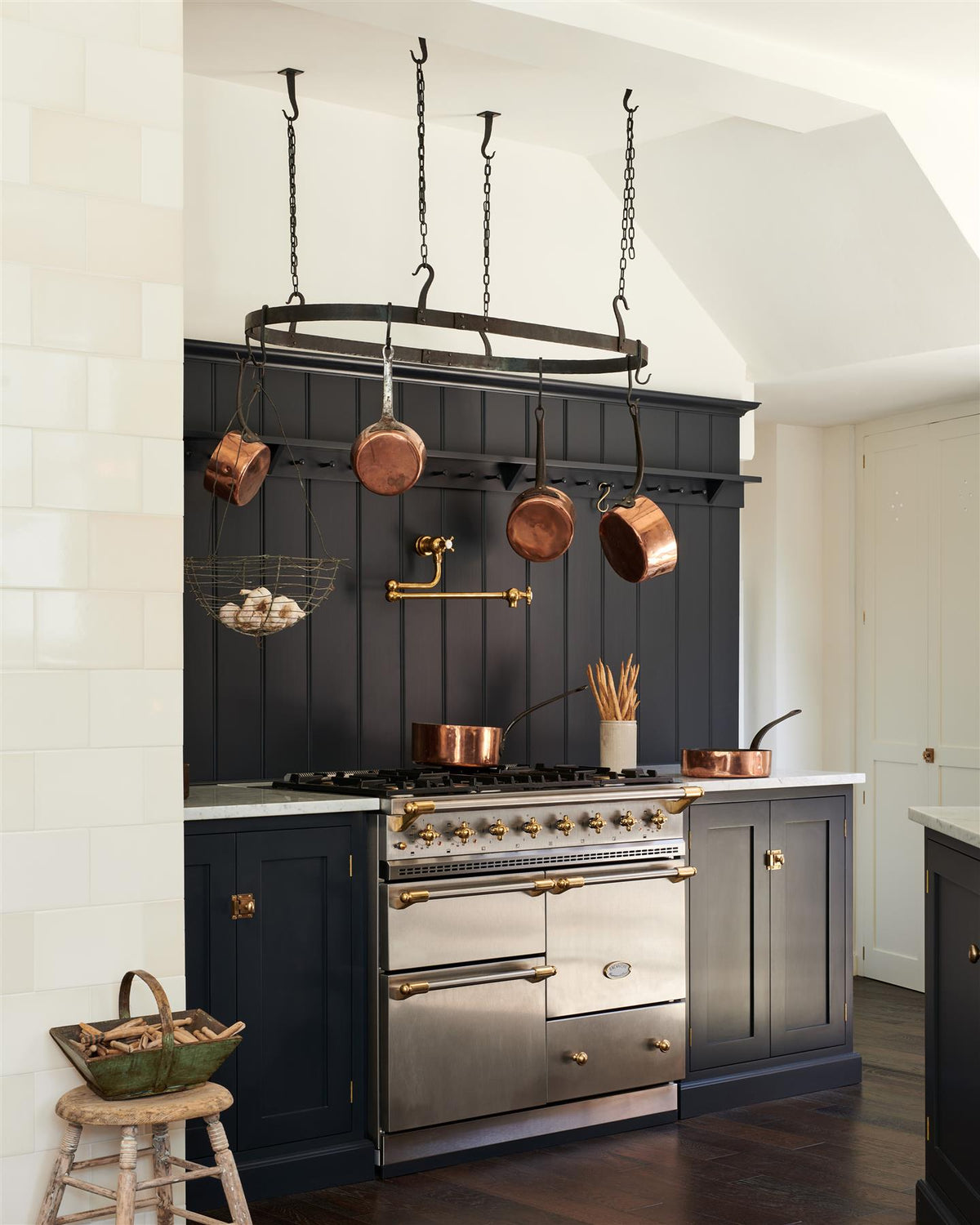 An oblique view of a simple Shaker-style kitchen with dark blue painted cabinetry and a stainless steel Lacanche range cooker with a large oval-shaped wrought iron pot hanger suspended from chains above. TEST