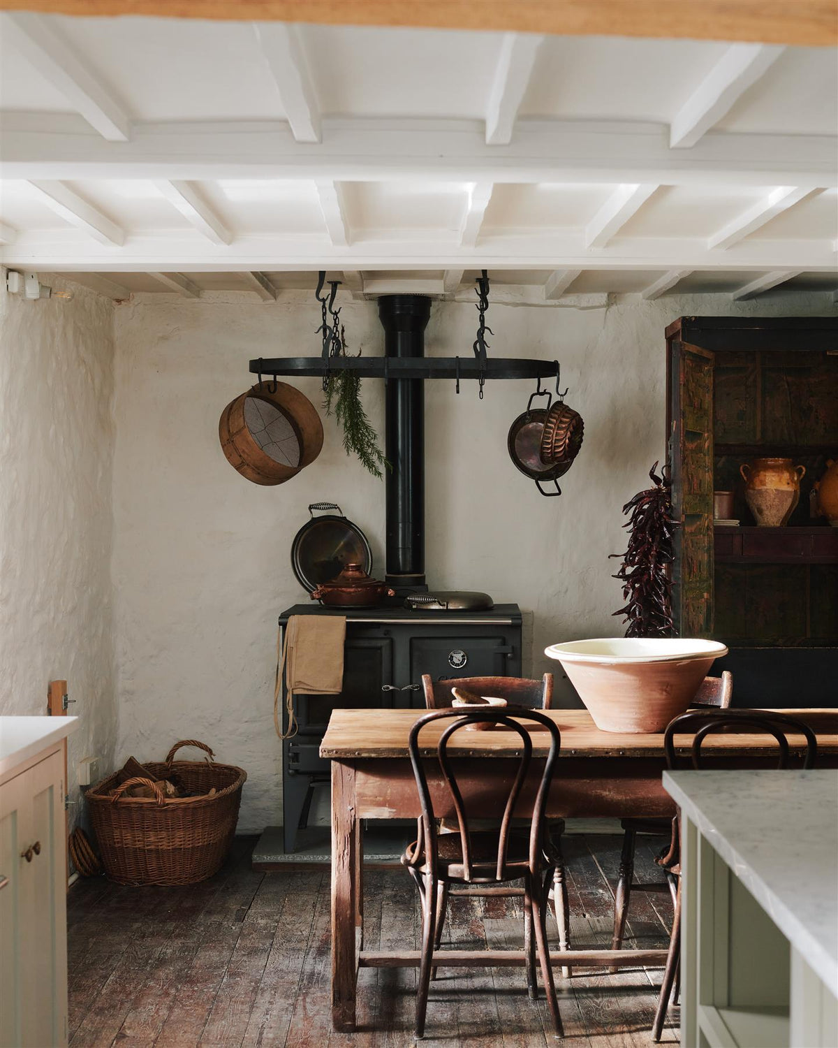 A cottage kitchen dining space with beamed ceilings, old worn floorboards, an antique pine table and chairs and a black Esse stove cooker with a large oval-shaped wrought iron pot hanger suspended from chains above. TEST