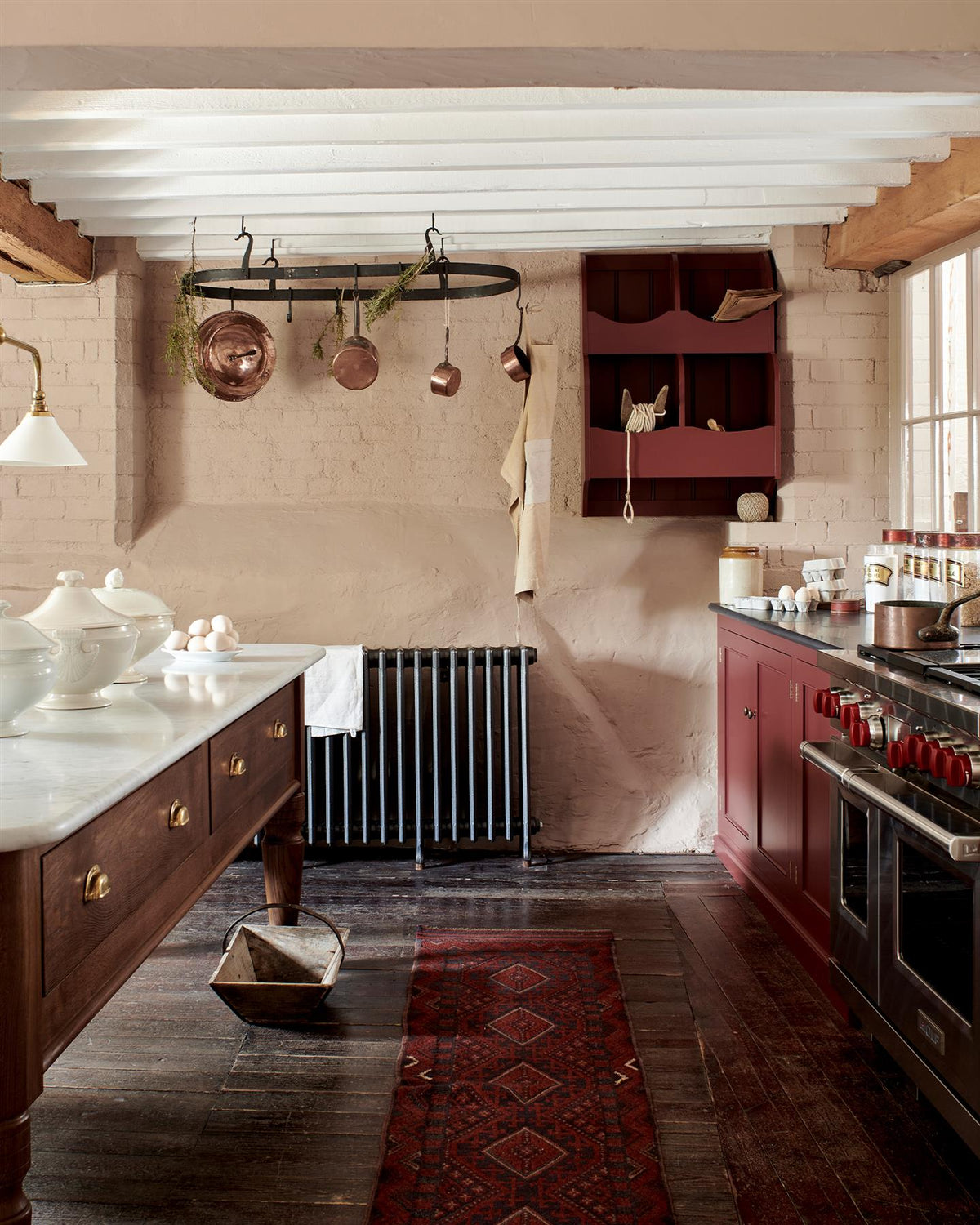 A Victorian-inspired country house kitchen with red painted cabinetry, worn floorboards, pink painted brick walls and a large oval-shaped wrought iron pot hanger suspended from a beamed ceiling via iron hooks. TEST