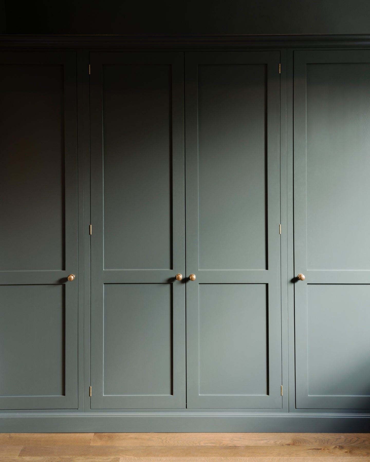 A straight-on view of dark sage green painted Shaker-style full height pantry cupboards, fitted with round doorknobs in a burnished 'Bella' brass finish.