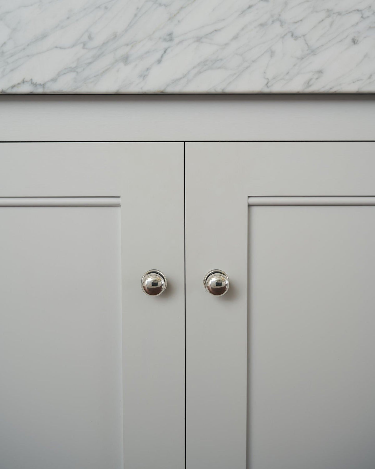 A straight-on view of a pair of round doorknobs with pressed backplates in a tarnished silver finish, fitted to white painted Georgian-style kitchen cupboard doors. TEST