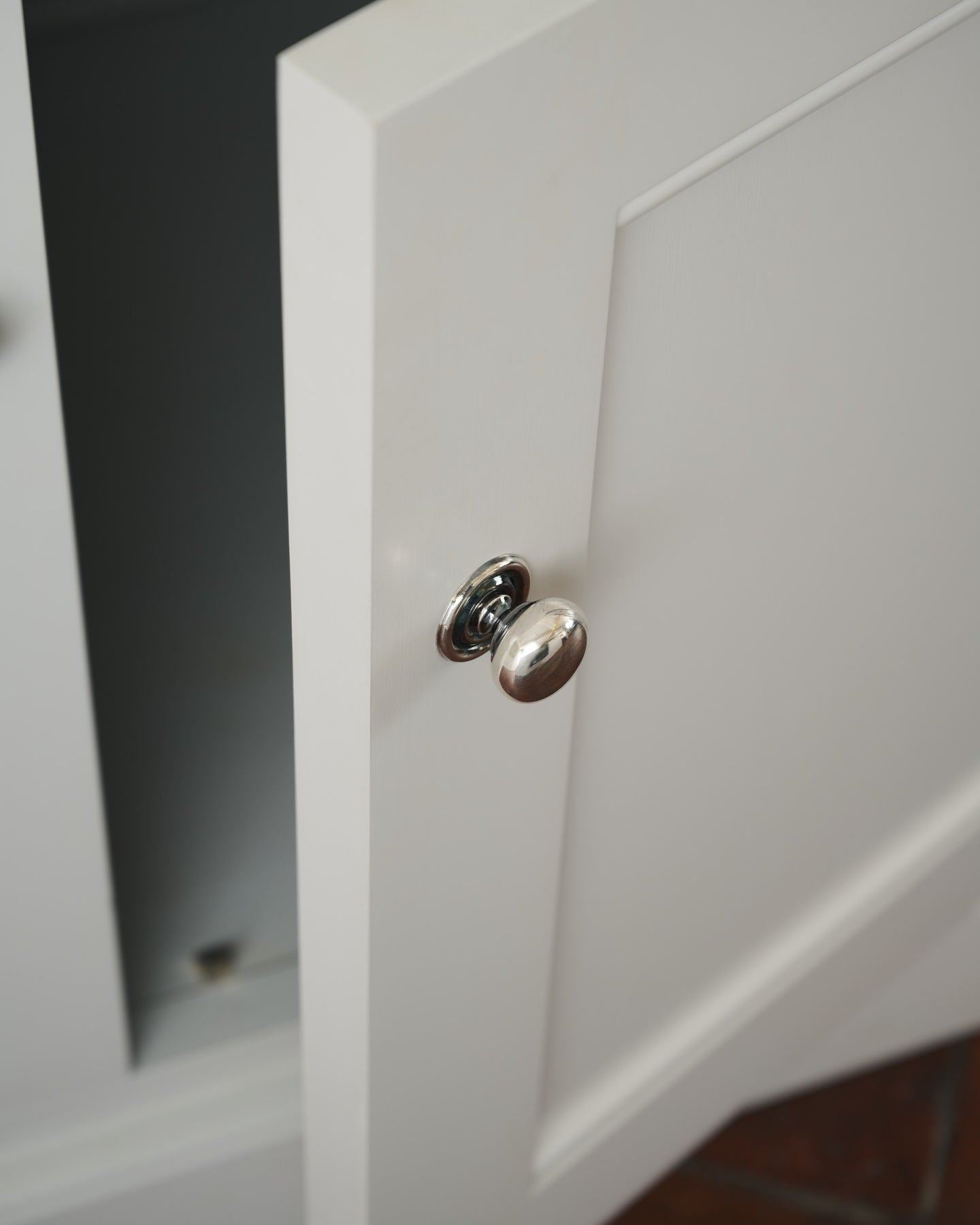 A round doorknob with a pressed backplate in a tarnished silver finish, fitted to an open white painted kitchen cupboard door.