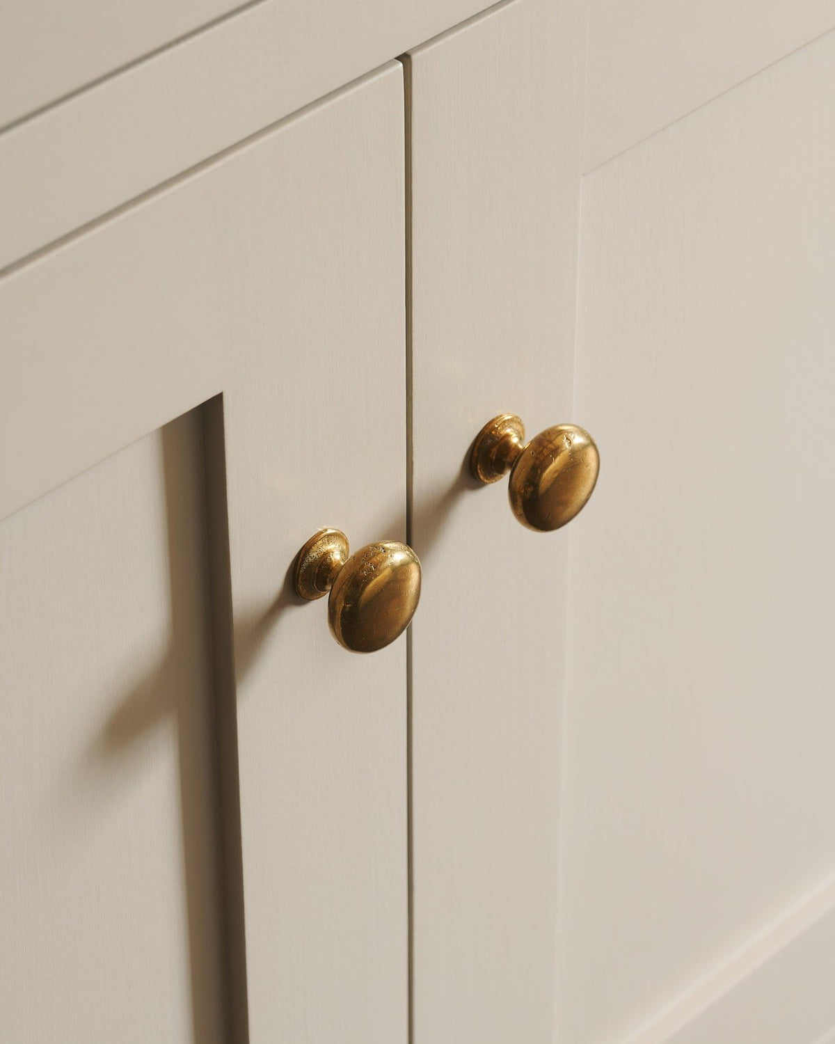 An oblique view of a pair of classic knobs in a warm burnished brass finish, fitted to a mushroom painted Shaker-style kitchen cupboard.|Medium shown TEST