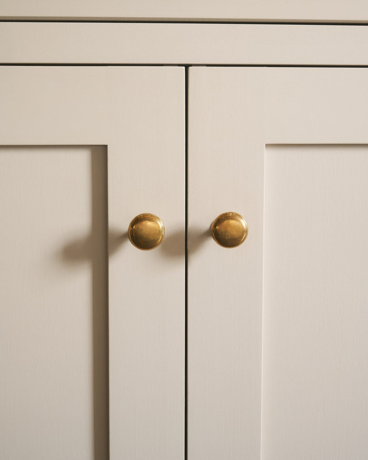 A straight-on view of a pair of classic knobs in a warm burnished brass finish, fitted to a mushroom painted Shaker-style kitchen cupboard. TEST