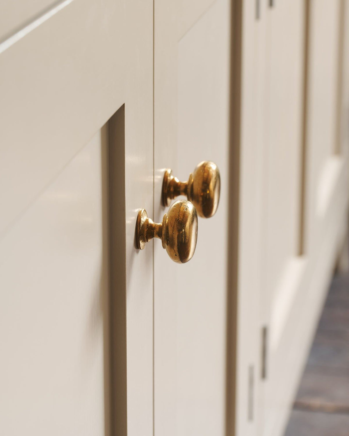 A close-up profile view of a pair of classic knobs in a warm burnished brass finish, fitted to a mushroom painted Shaker-style kitchen cupboard.|Medium shown TEST