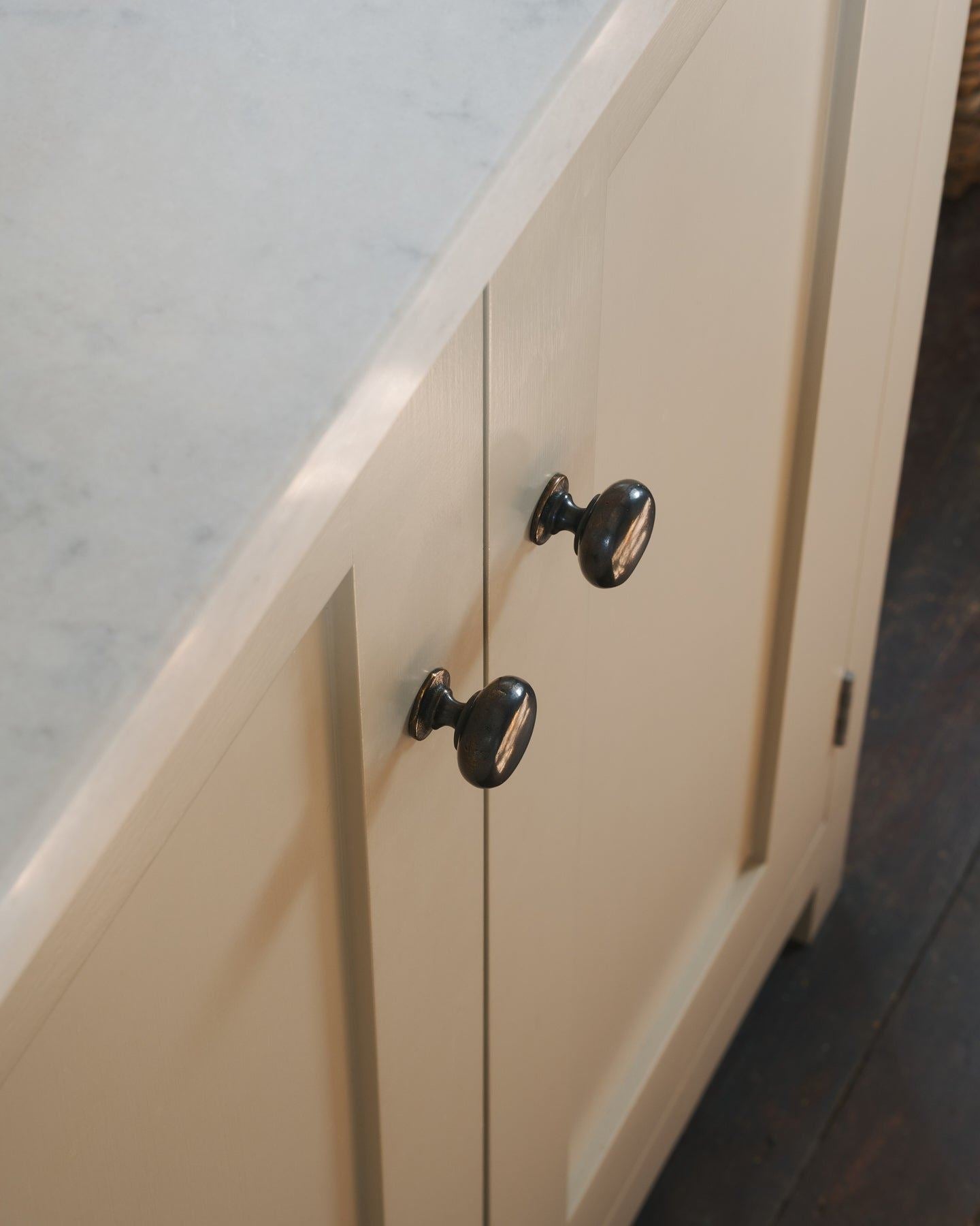 A close-up oblique view of a pair of classic knobs in a dark oxidised bronze finish, fitted to a mushroom painted Shaker-style kitchen cupboard.