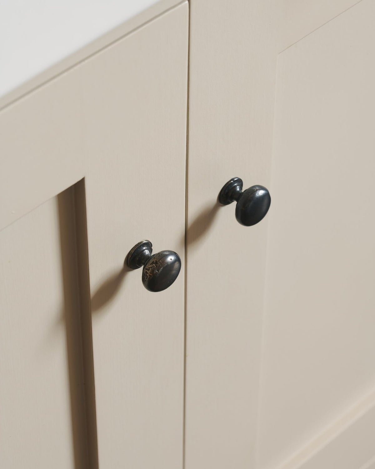 An oblique view of a pair of classic knobs in a dark oxidised bronze finish, fitted to a mushroom painted Shaker-style kitchen cupboard. TEST