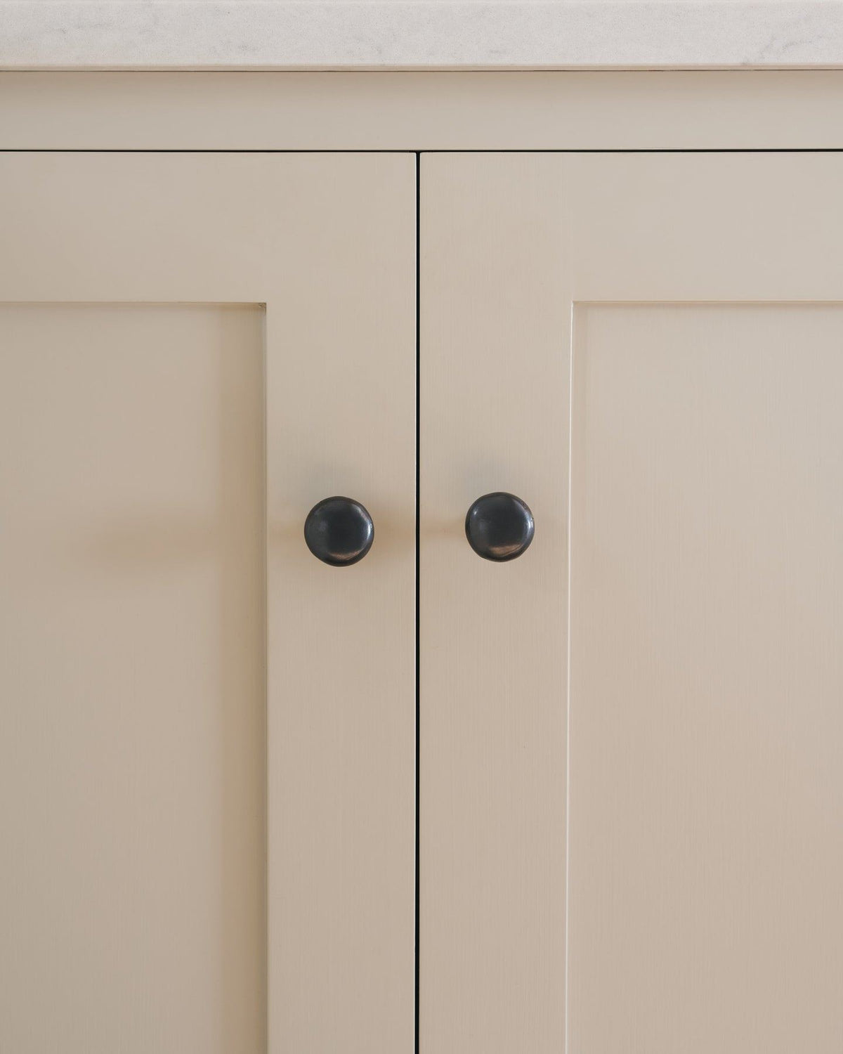 A straight-on view of a pair of classic knobs in a dark oxidised bronze finish, fitted to a mushroom painted Shaker-style kitchen cupboard. TEST