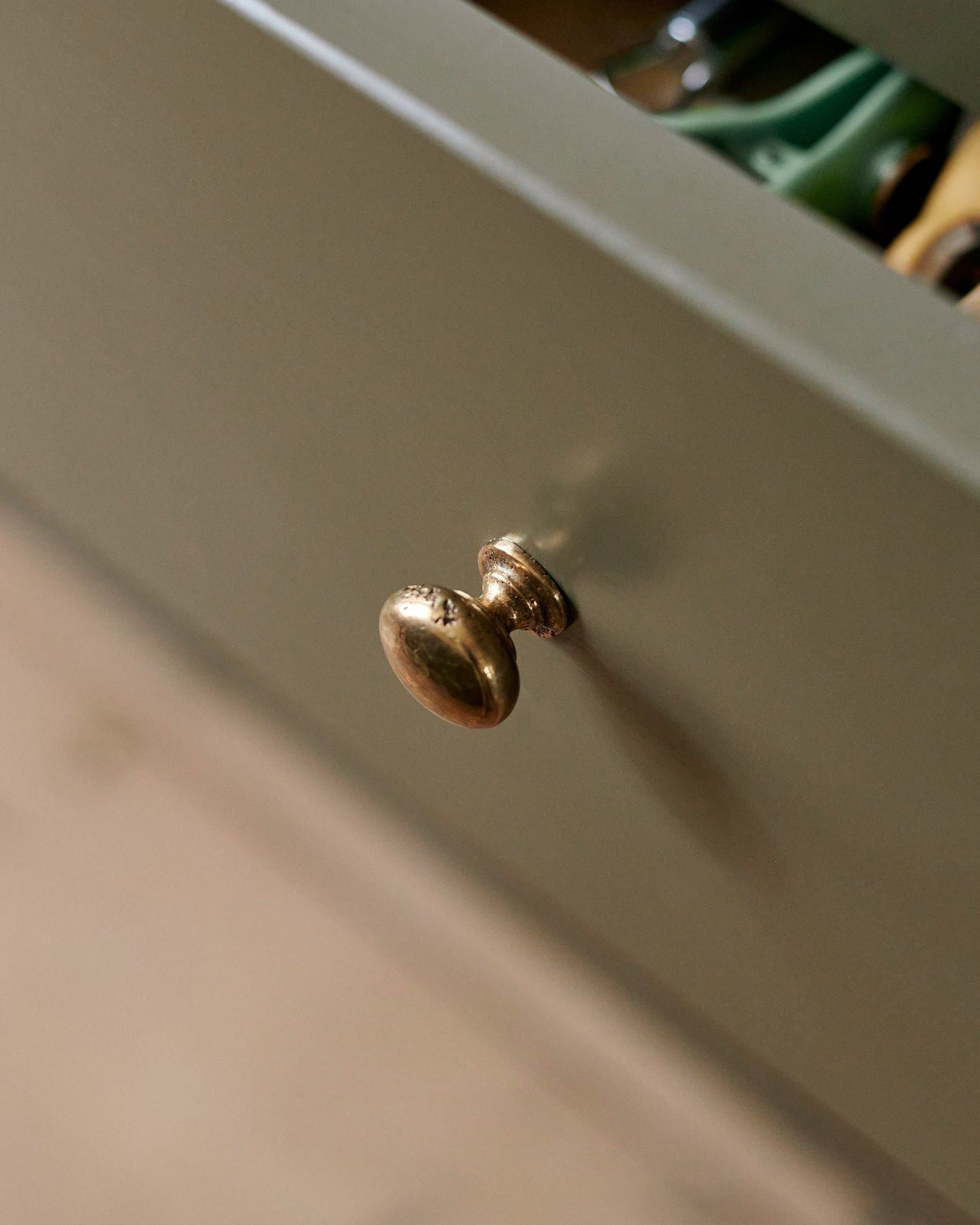 A close-up oblique view of a classic knob in a bright polished bronze finish, fitted to a mossy green painted drawer front.