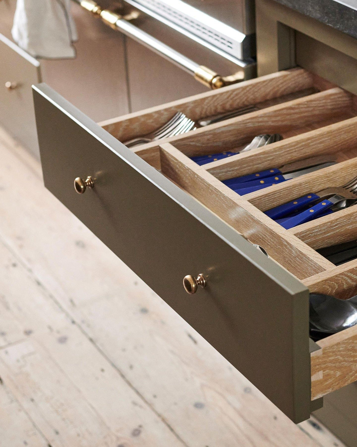An oblique view of a mossy green painted open cupboard drawer with classic knobs in a bright polished bronze finish and an oak cutlery tray full of utensils.|Medium shown TEST