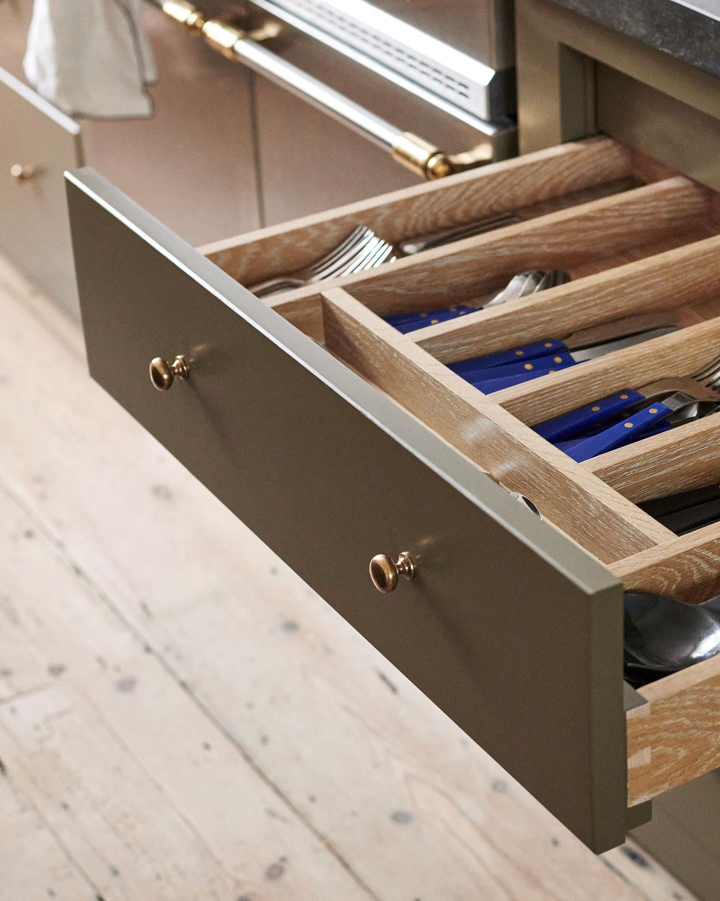 An oblique view of a mossy green painted open cupboard drawer with classic knobs in a bright polished bronze finish and an oak cutlery tray full of utensils.