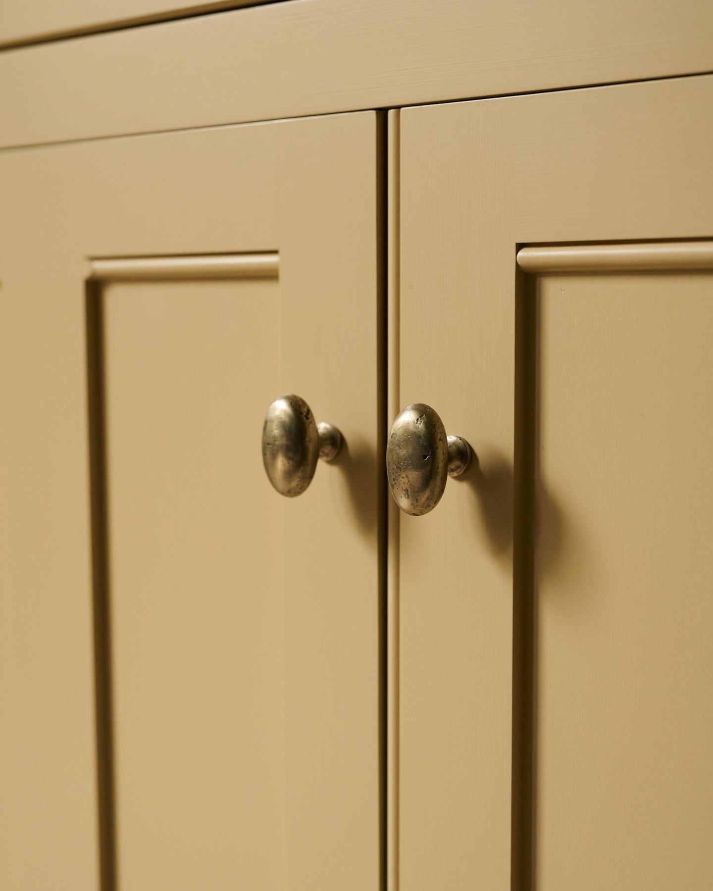 An oblique view of a pair of oval-shaped knobs in an aged brass finish, fitted to a yellow painted Georgian-style kitchen cupboard.