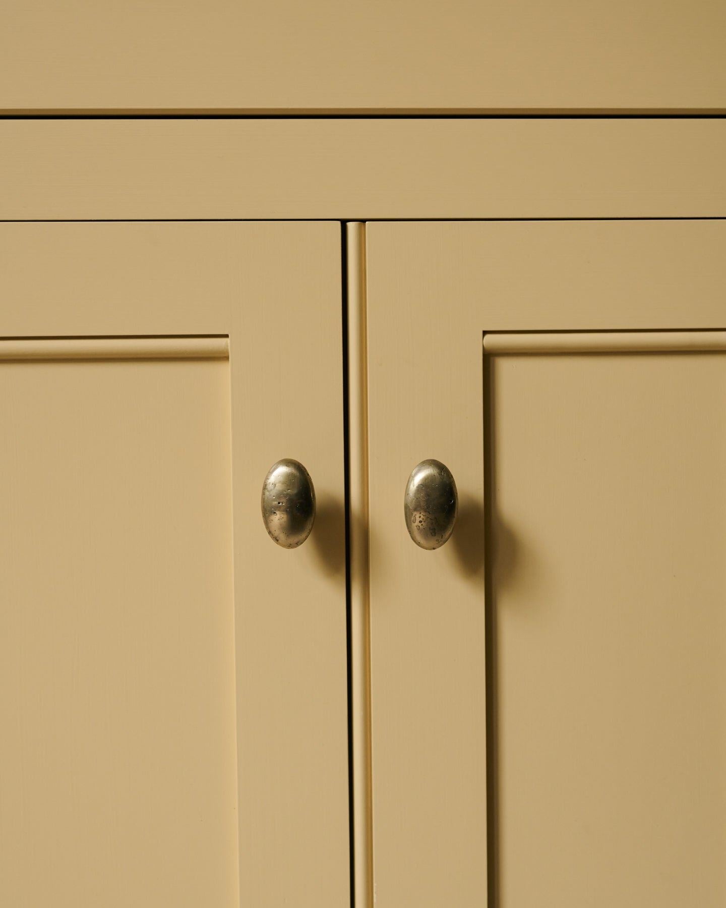 A straight-on view of a pair of oval-shaped knobs in an aged brass finish, fitted to a yellow painted Georgian-style kitchen cupboard.