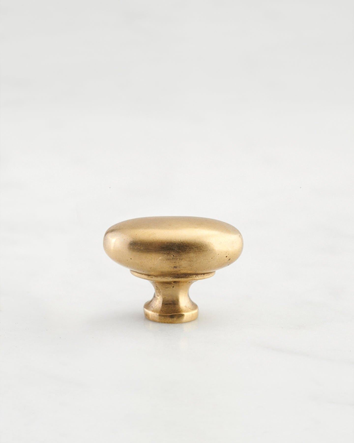 A 'Petite' oval-shaped knob for a kitchen cupboard or drawer with an aged brass finish and unique dented textures that makes it look like a beloved, well aged brass from centuries ago.