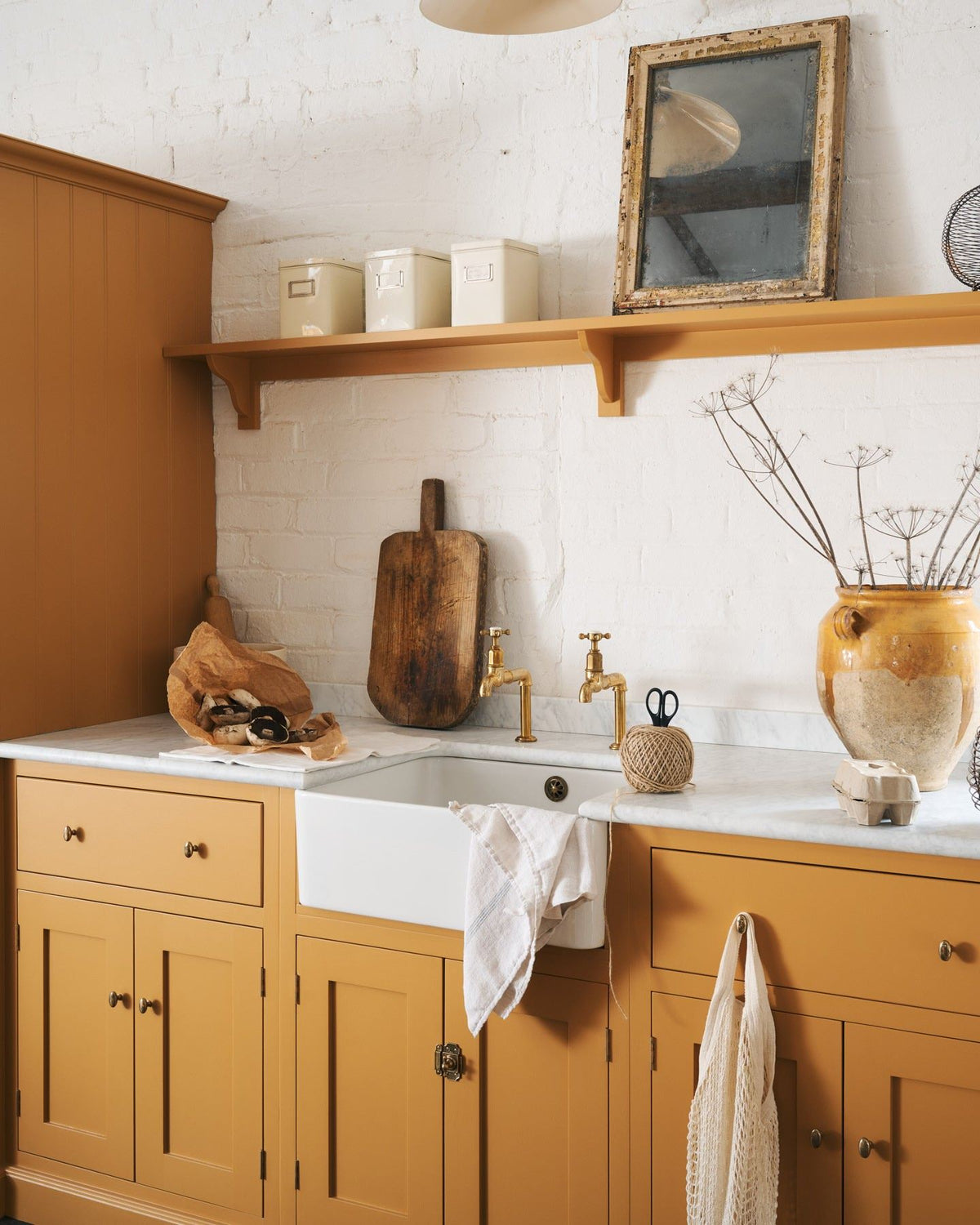 An oblique view of a yellow painted Shaker-style kitchen sink cupboard with small oval-shaped knobs in a warm burnished brass finish.|Heirloom Brass Small shown TEST