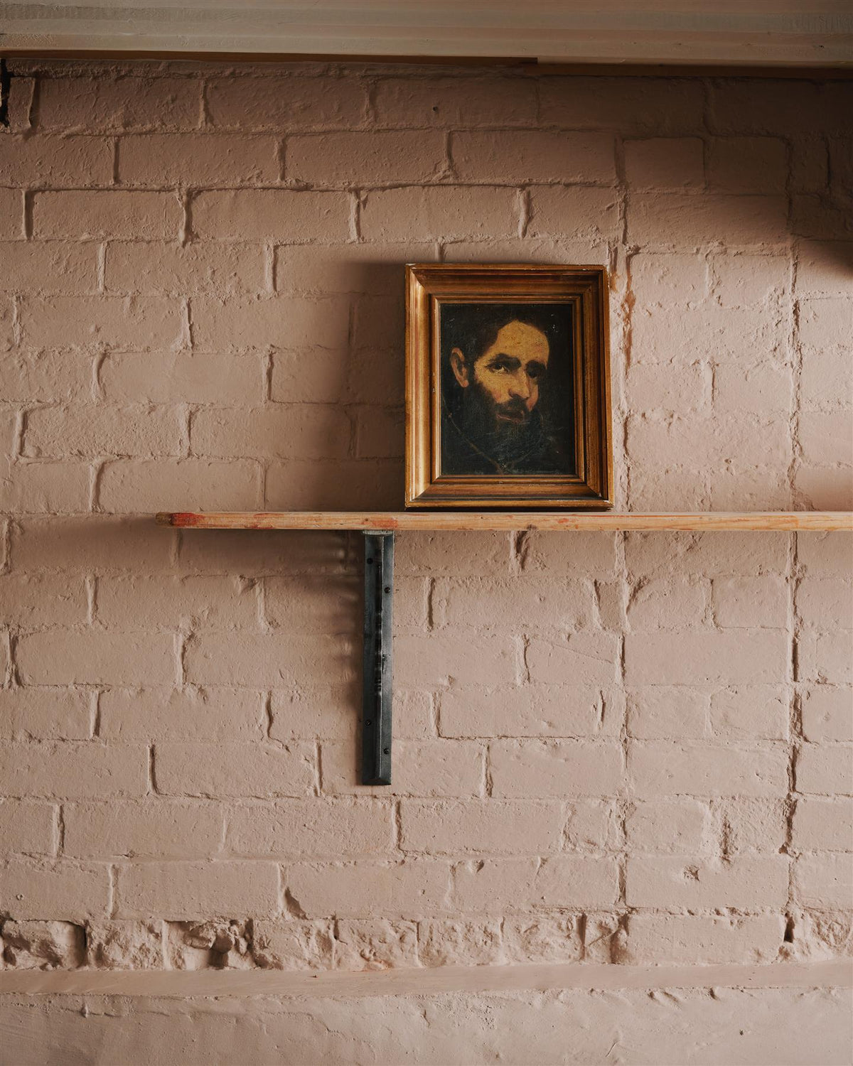 A straight-on view of a large Victorian-inspired ornate cast iron shelf bracket holding a deep pine shelf to a pink colour brick wall. TEST