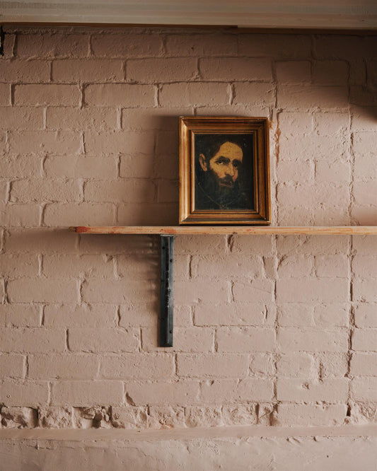 A straight-on view of a large Victorian-inspired ornate cast iron shelf bracket holding a deep pine shelf to a pink colour brick wall.