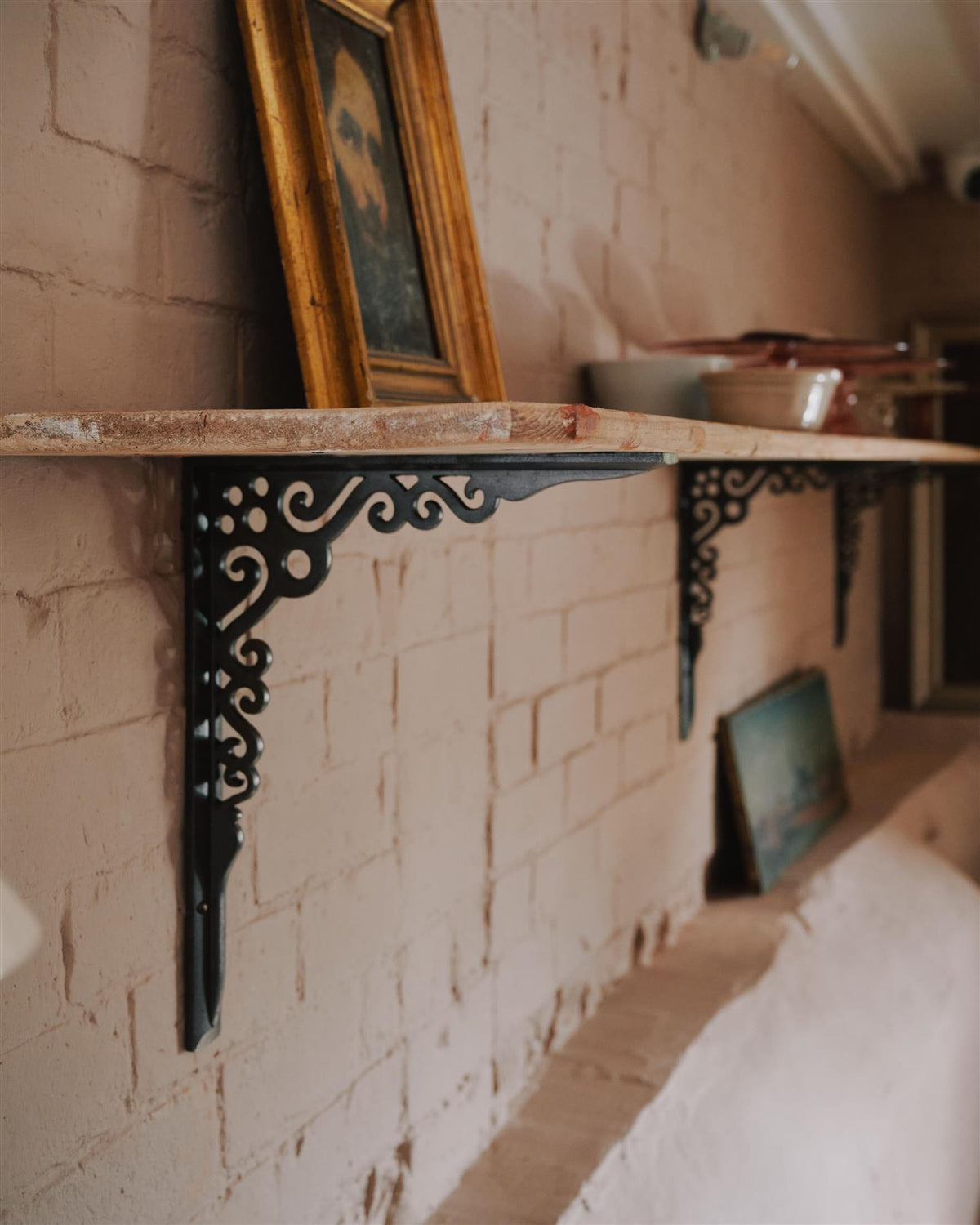 An oblique view of a large Victorian-inspired ornate cast iron shelf bracket holding a deep pine shelf to a pink colour brick wall. TEST