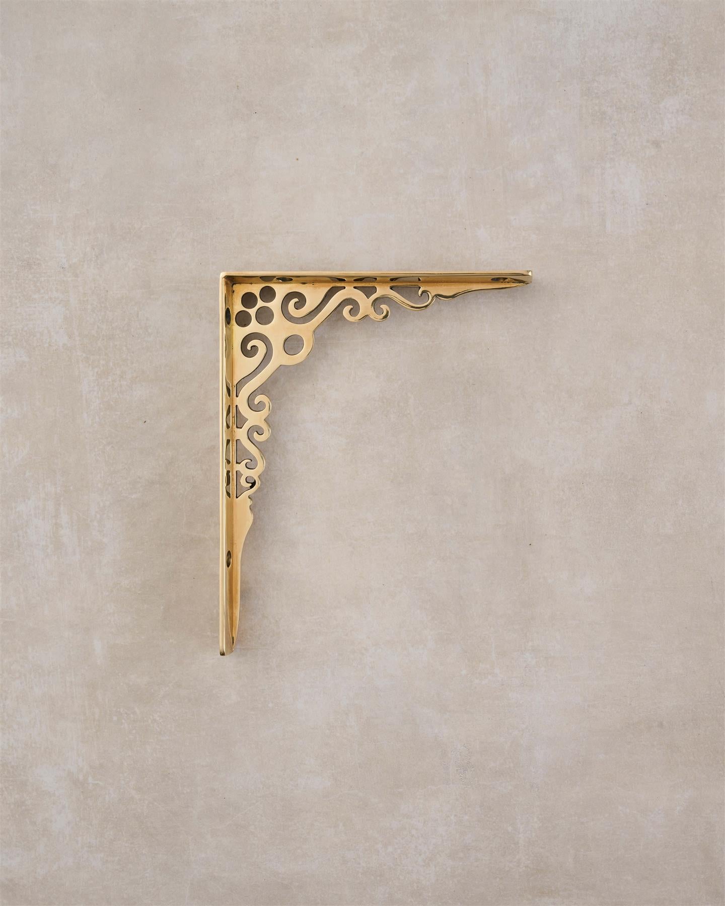 A side profile view of a small Victorian-inspired ornate shelf bracket in a warm polished brass finish.