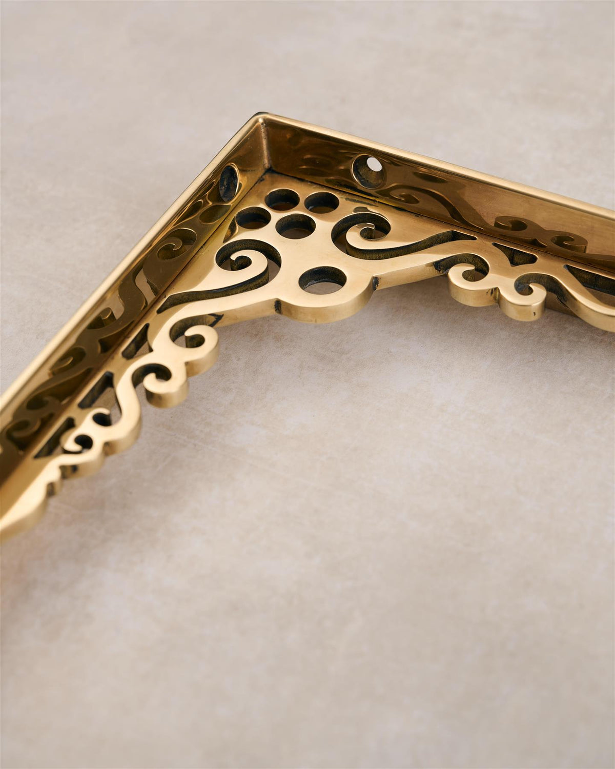 A close-up view of the decorative cutout patterns on a small Victorian-inspired ornate polished brass shelf bracket. TEST