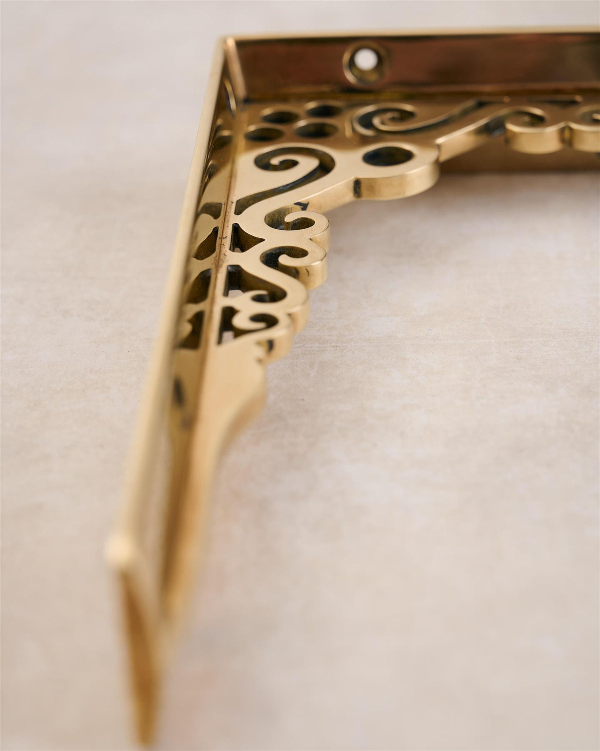 A close-up view of the decorative cutout patterns on a small Victorian-inspired ornate polished brass shelf bracket. TEST