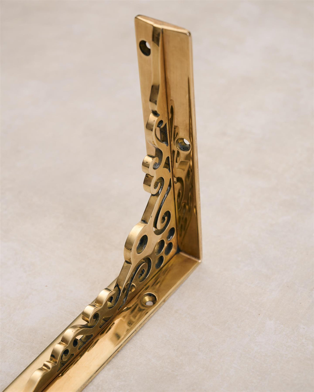 A close-up oblique view of a small Victorian-inspired ornate shelf bracket in a warm polished brass finish. TEST