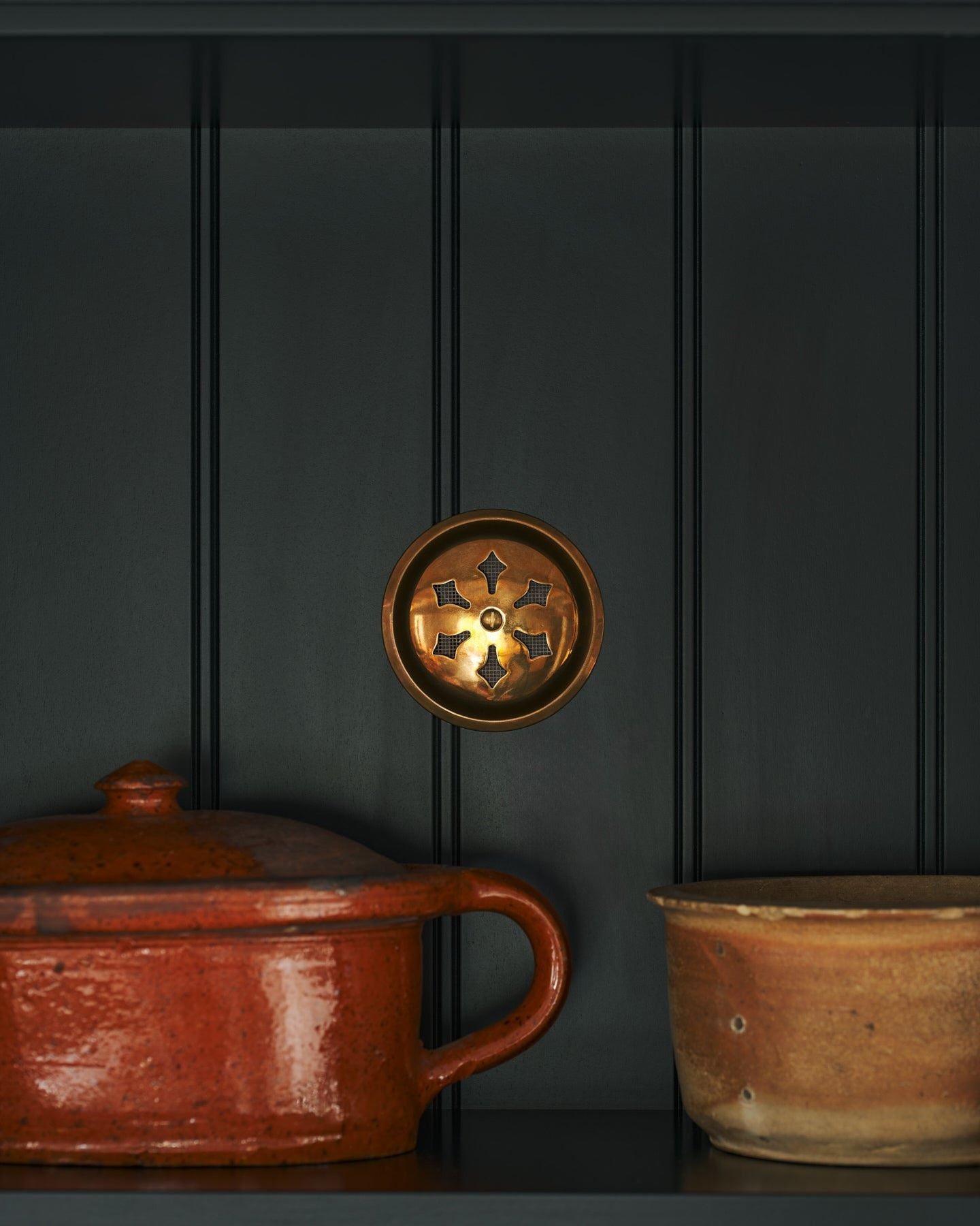 A Victorian-inspired circular brass ventilation grille with traditional decorative cutouts fitted to a black painted tongue and groove-style cupboard back panel.