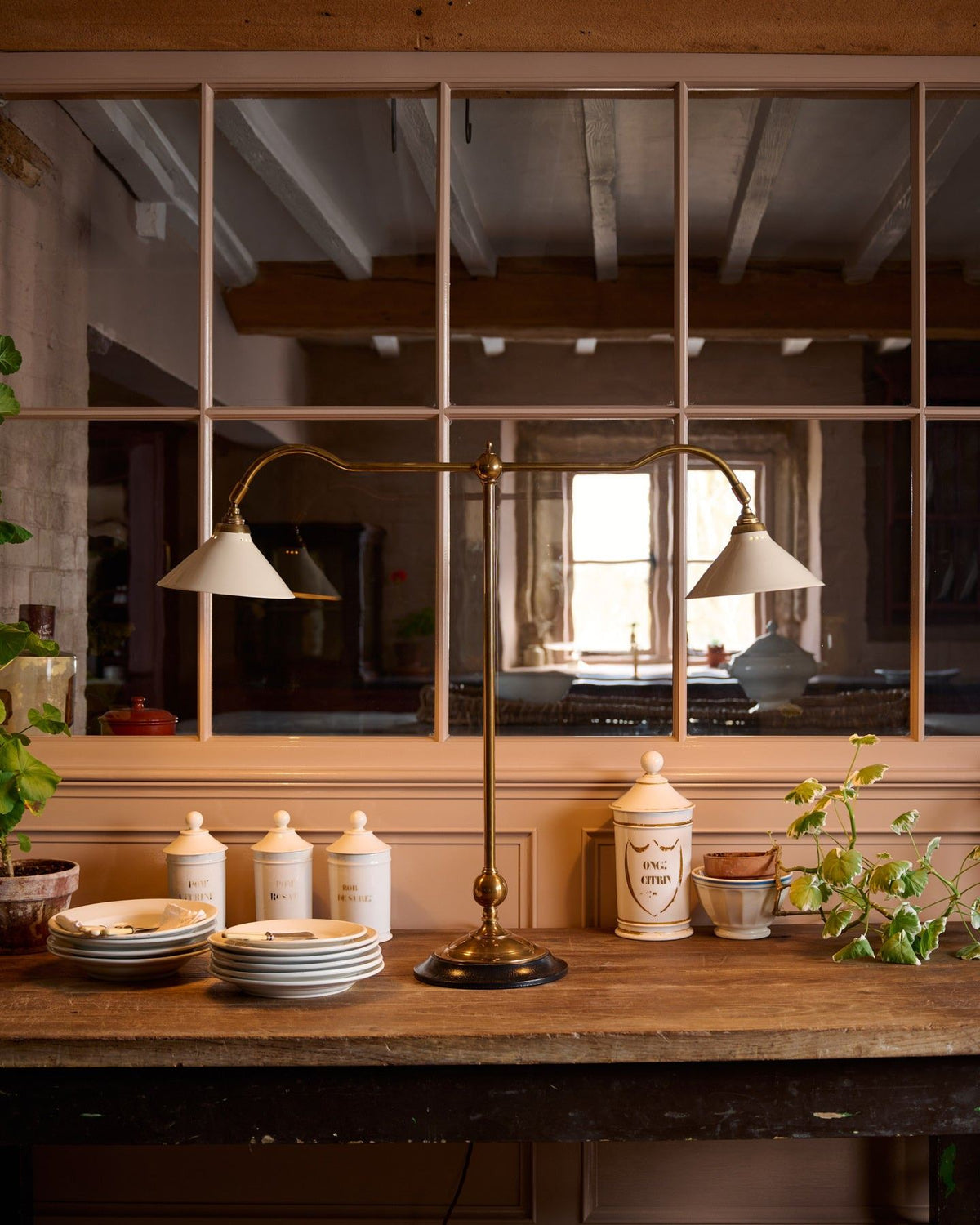 A straight-on view of a Victorian-inspired desk lamp with a solid brass foot, stand, two curved arms and two small cream glazed ceramic light shades on a table in front of a glazed partition. TEST