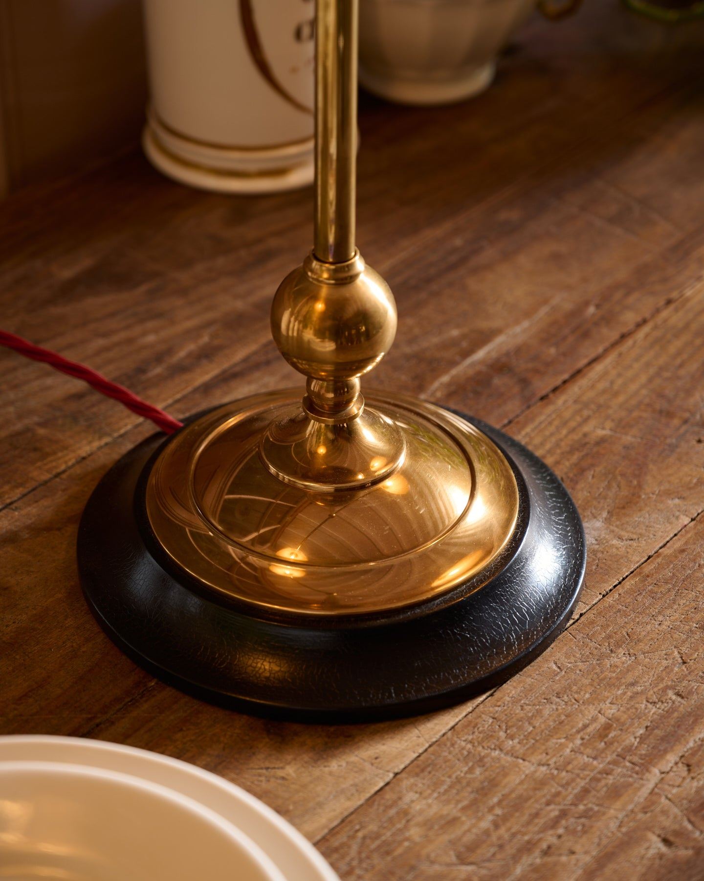 A close-up view of a solid brass desk lamp foot with a dark hardwood base and a red twisted fabric flex cord leading out from one side.