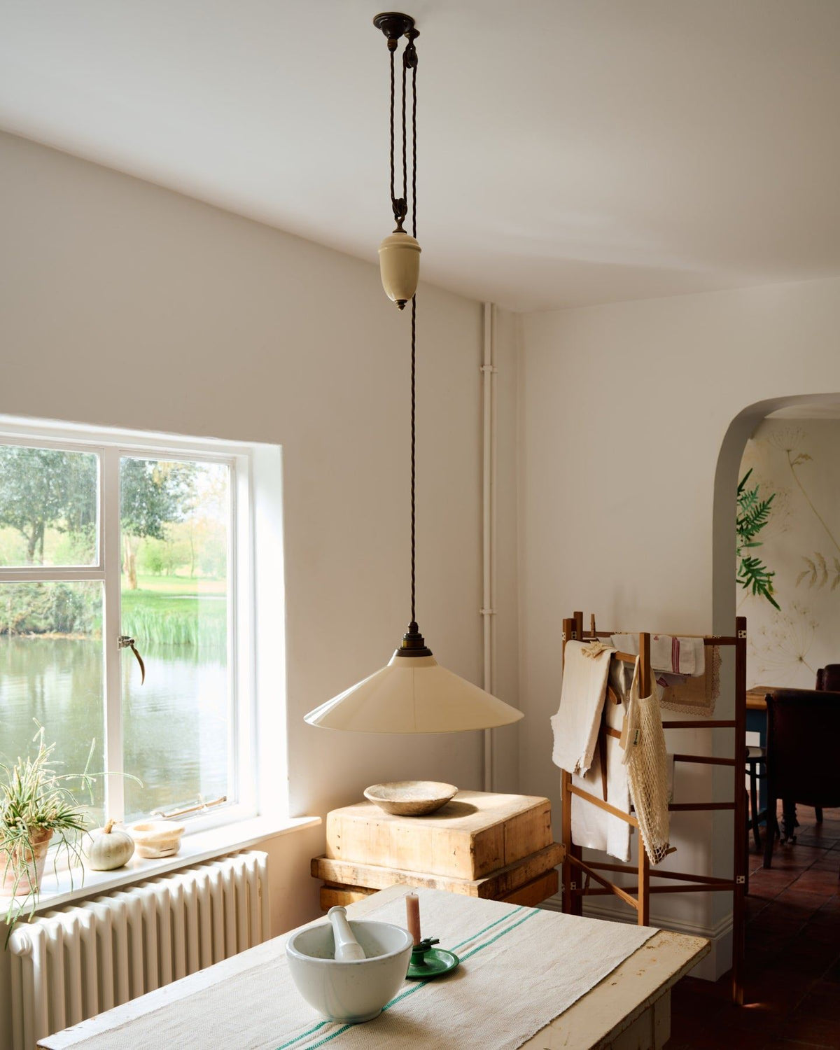 An oblique view of a rise and fall light with a large cream glazed ceramic shade, aged brass fittings and brown twisted fabric flex cord hanging in a white room above an antique pine table. TEST