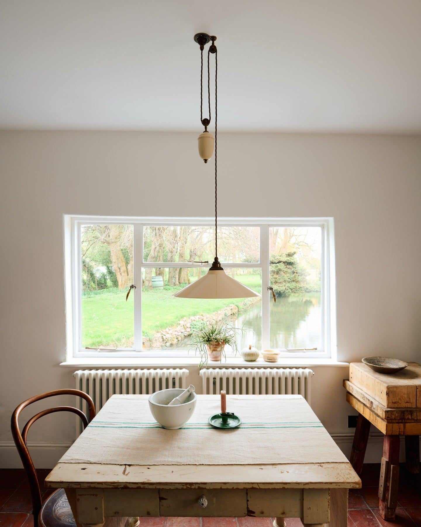 A straight-on view of a rise and fall light with a large cream glazed ceramic shade, aged brass fittings and brown twisted fabric flex cord hanging in a white room above an antique pine table.