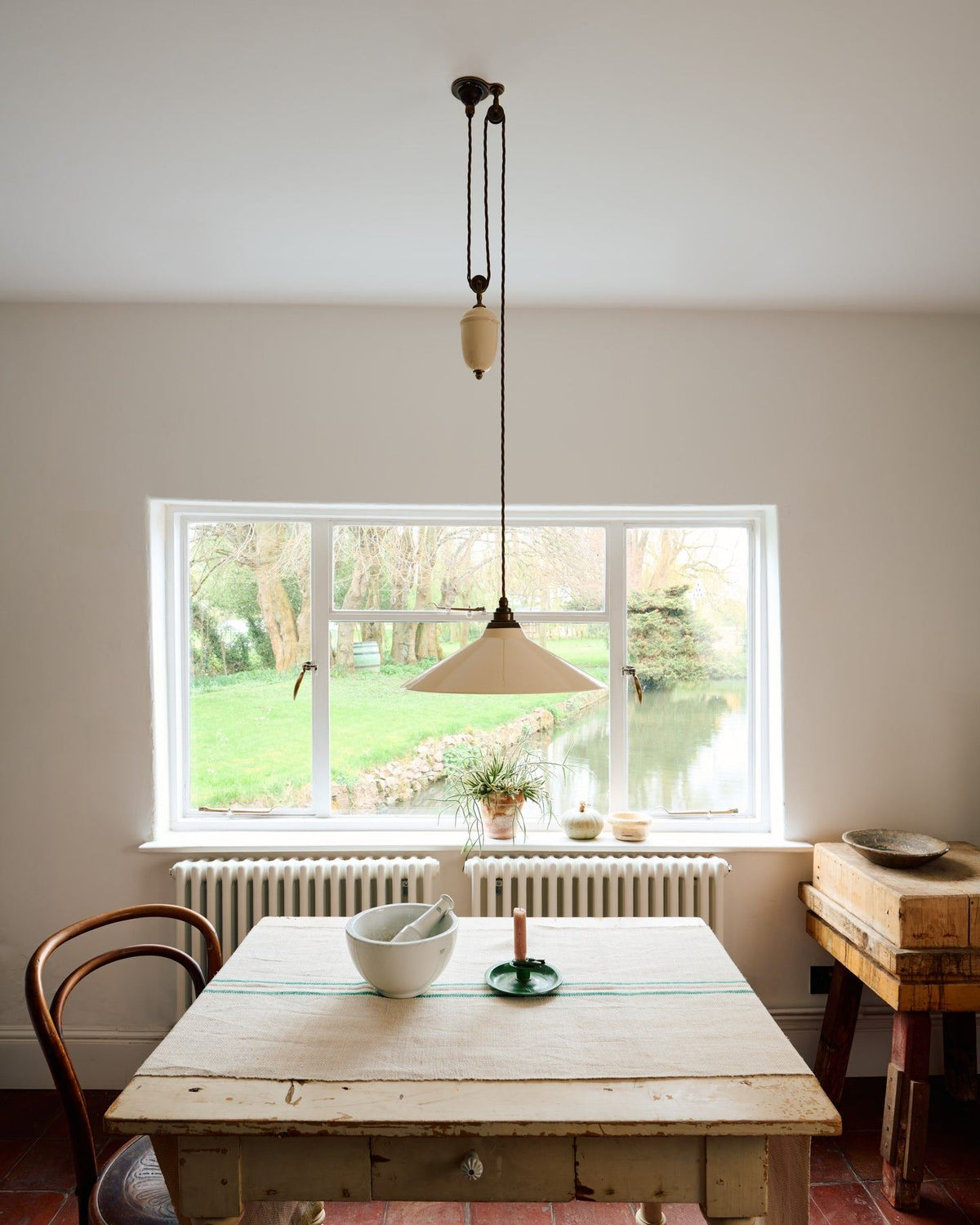 A straight-on view of a rise and fall light with a large cream glazed ceramic shade, aged brass fittings and brown twisted fabric flex cord hanging in a white room above an antique pine table. TEST