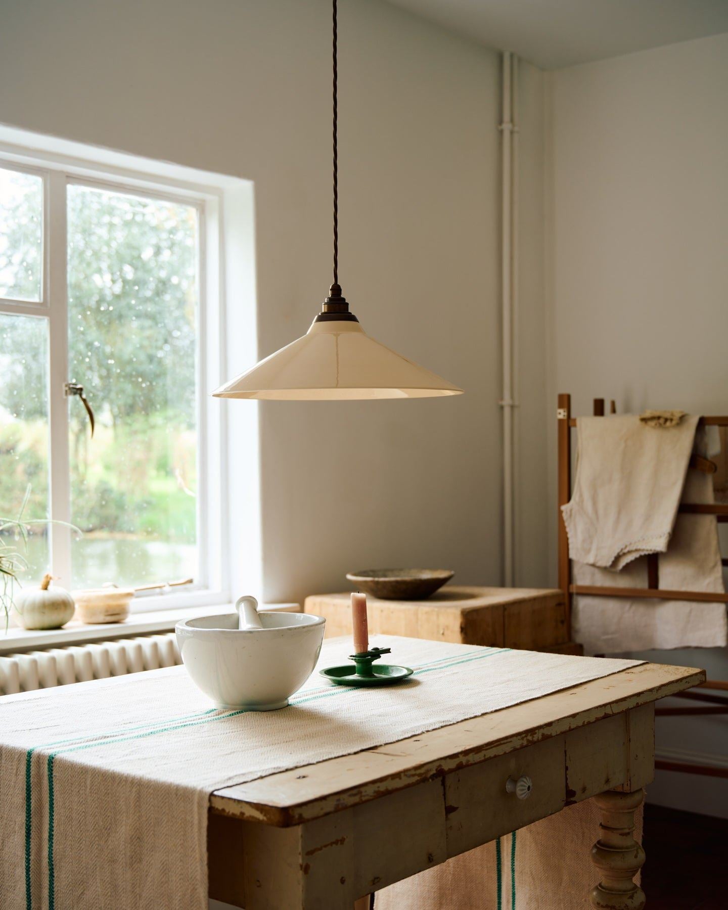 A close-up oblique view of a rise and fall light with a large cream glazed ceramic shade, aged brass fittings and brown twisted fabric flex cord hanging in a white room above an antique pine table.