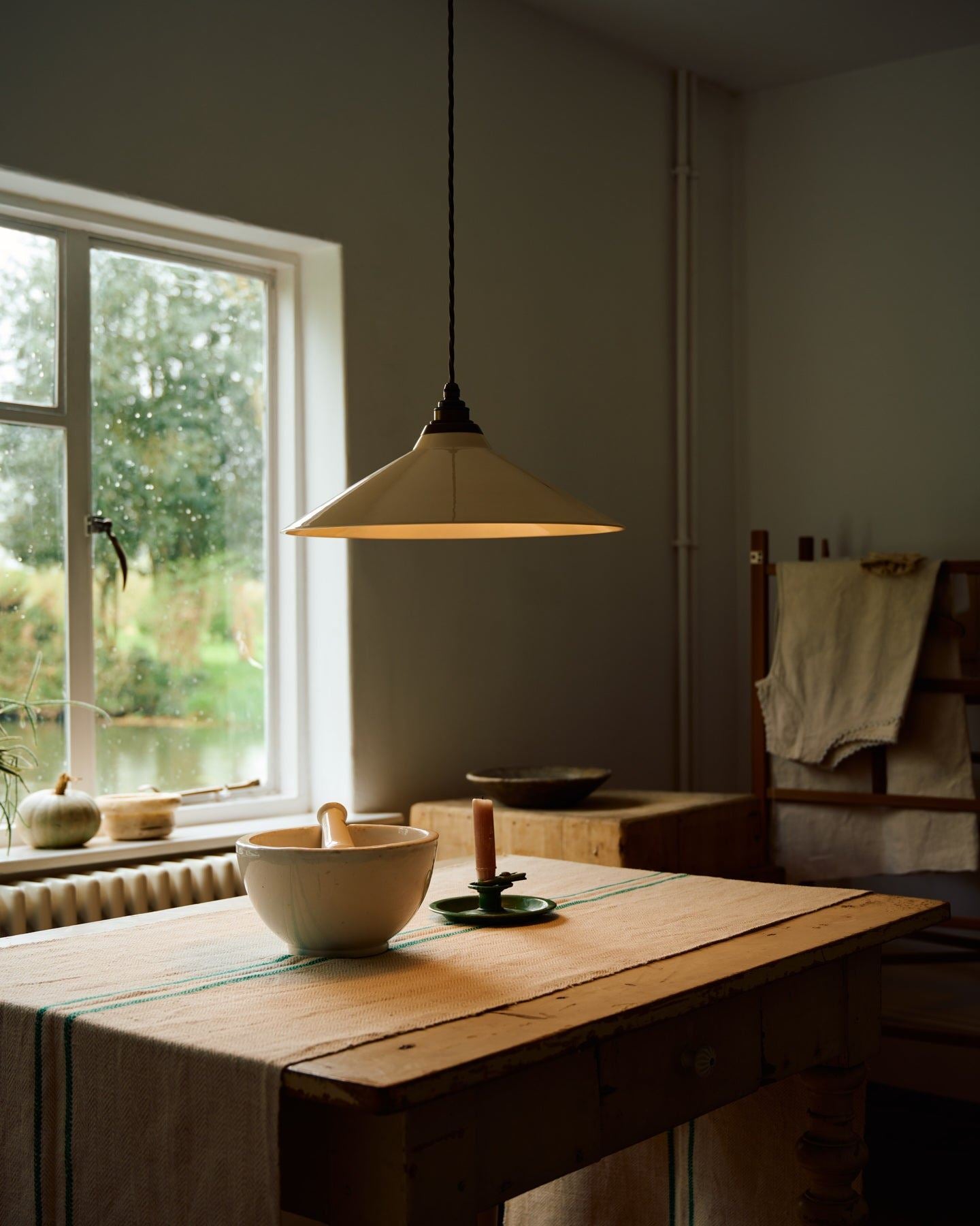 A close-up oblique view of a rise and fall light, turned on, with a large cream glazed ceramic shade, aged brass fittings and brown twisted fabric flex cord hanging in a white room above an antique pine table.