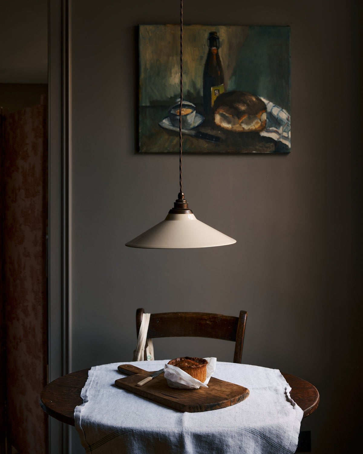 A straight-on view of a rise and fall light with a small cream glazed ceramic shade, aged brass fittings and brown twisted fabric flex cord hanging in a dark room over a small round wooden table with a linen tablecloth and fresh bread. TEST