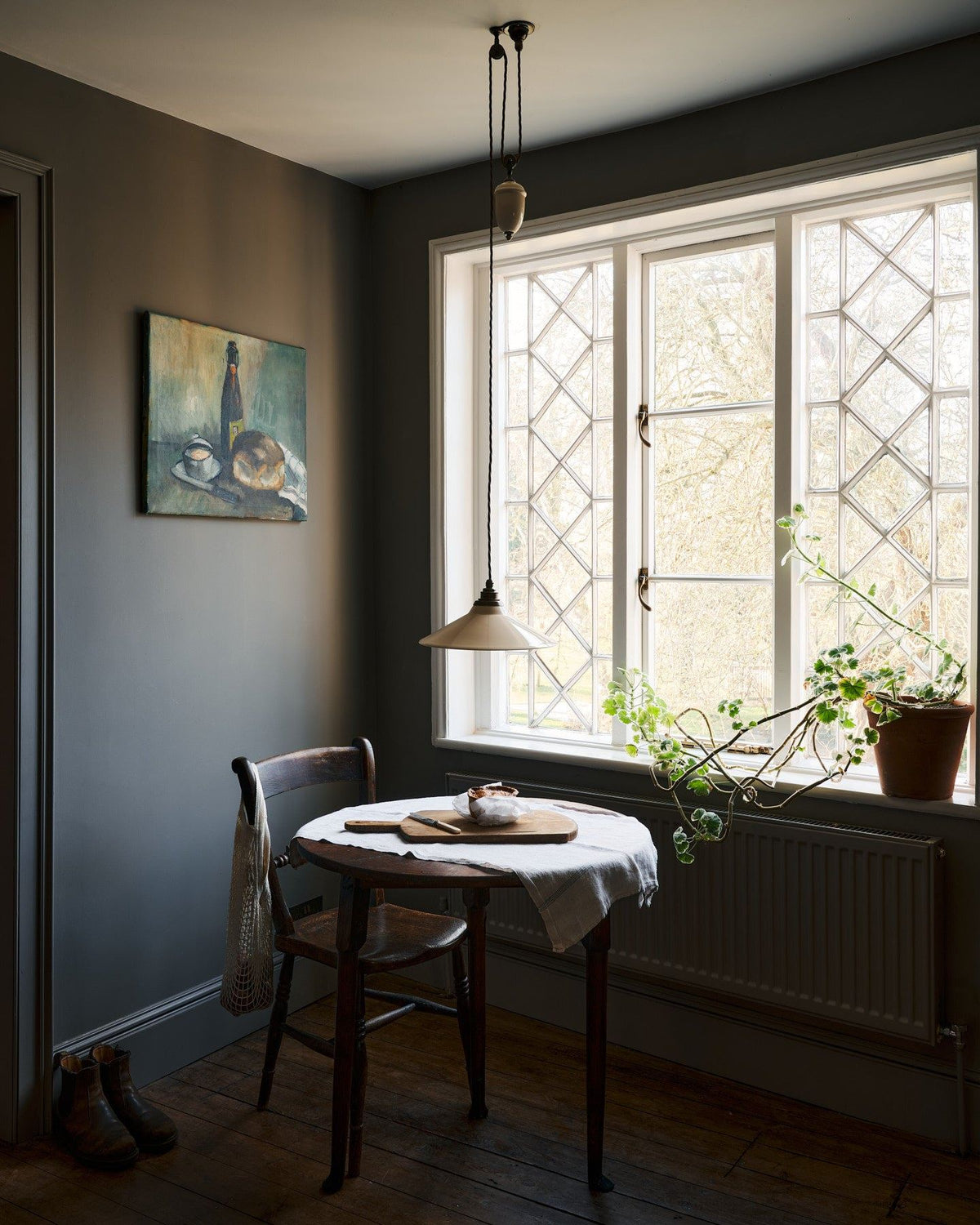 An oblique view of a rise and fall light with a small cream glazed ceramic shade, aged brass fittings and brown twisted fabric flex cord hanging in a dark room by a large Victorian leaded window. TEST
